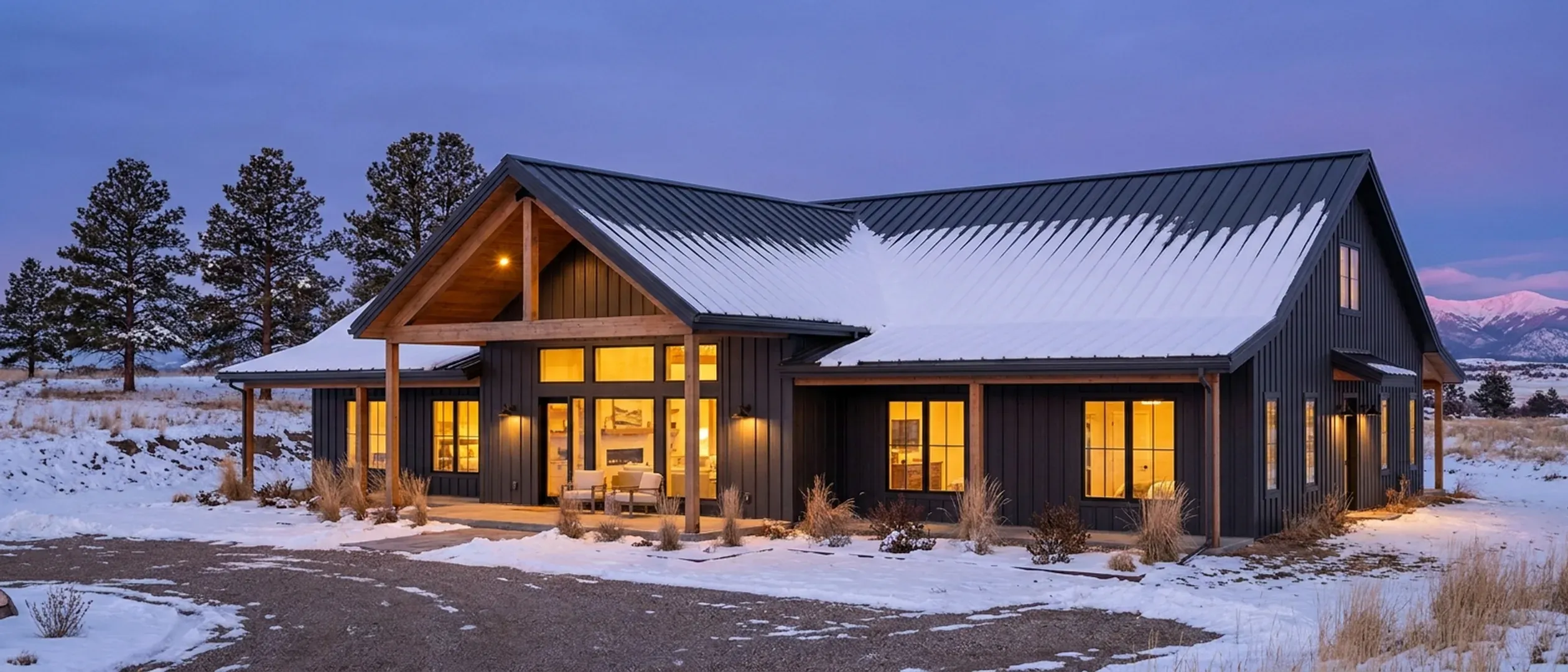 A barndominium at dusk in winter, snow on its standing seam roof and warm light in every window, with distant snow-capped mountains under a deep blue sky.